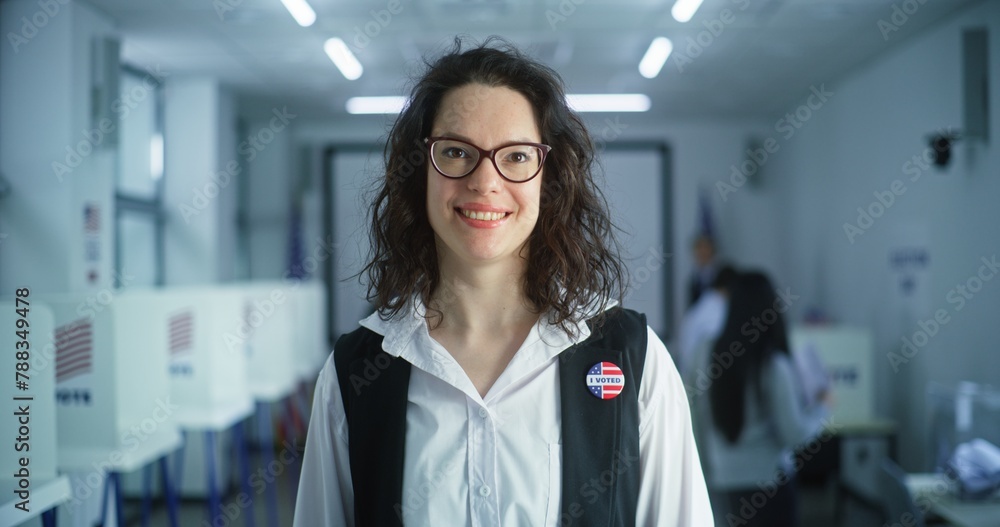 Fototapeta premium Portrait of Caucasian woman, United States of America elections voter. Woman with badge stands in polling station, poses, smiles, looks at camera. Background with voting booths. Concept of civic duty.