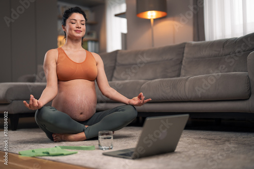 A focused mid adult pregnant lady meditating in her living room in front of a laptop photo