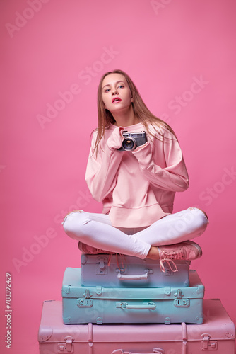 Travel concept. Studio portrait of pretty young woman holding camera sitting on baggage valises. Pink background. © luengo_ua