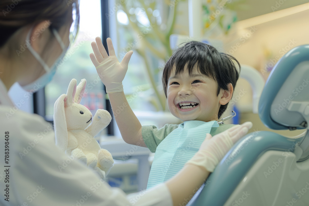 A Japanese boy, holding a toy rabbit, sits in the dental chair and happily high-fives the nurse during the doctor's appointment.