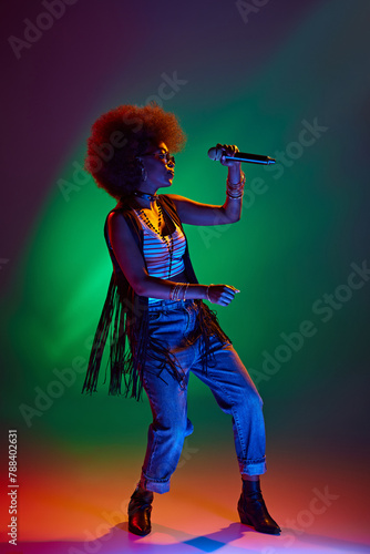 Portrait of young African-American woman dressed retro attire engaging in song in neon light against gradient background. Concept of art, music, hobby, classical music and modern lifestyle. Ad