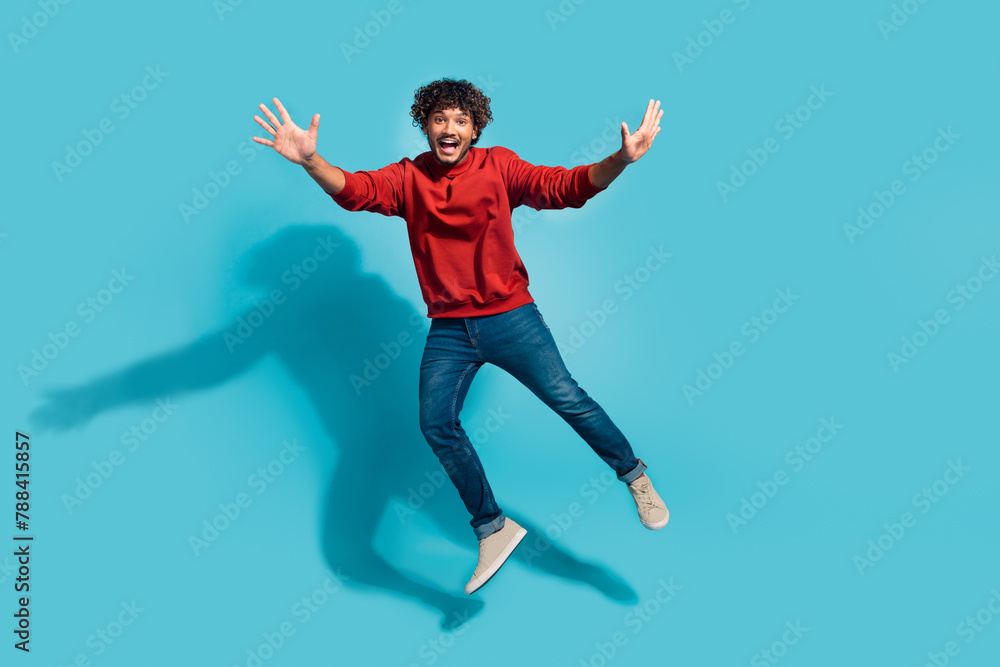 Full length body size photo of overjoyed curly hair hispanic guy raised arms up having fun jumping isolated on blue color background