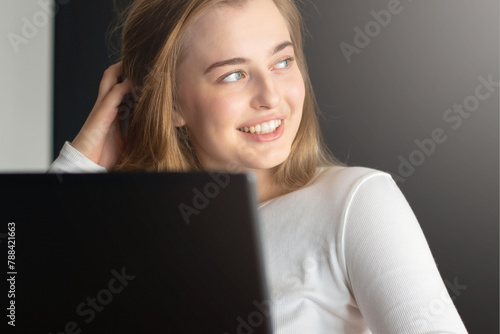 A beautiful young girl with blue eyes and long blond hair smiling out the window near the laptop on the table.