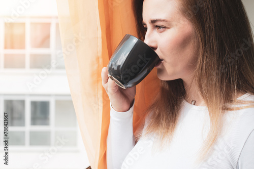 A blonde girl in profile is standing by the window drinking morning coffee. The young girl is drinking coffee thoughtfully. 