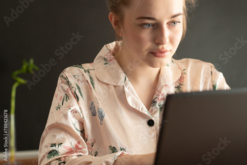 A blonde girl is working at home laptop in pink pajamas in the kitchen. Remote work at home.