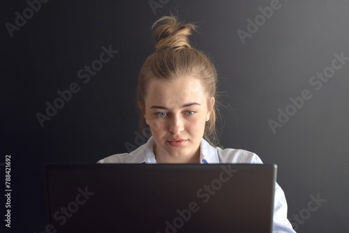 A close-up of a girl with blonde hair in a bundle and a light shirt is working with her laptop. 