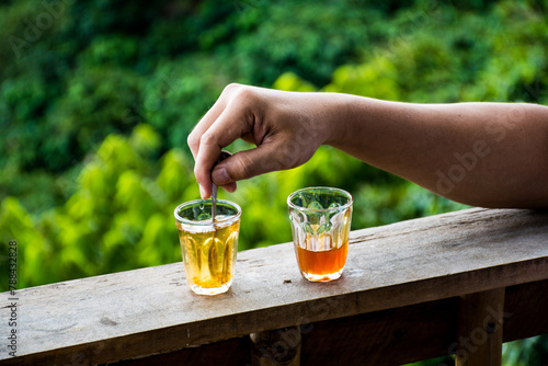 A hand is stirring tea in a glass cup, on a wooden bar, green-leaf background