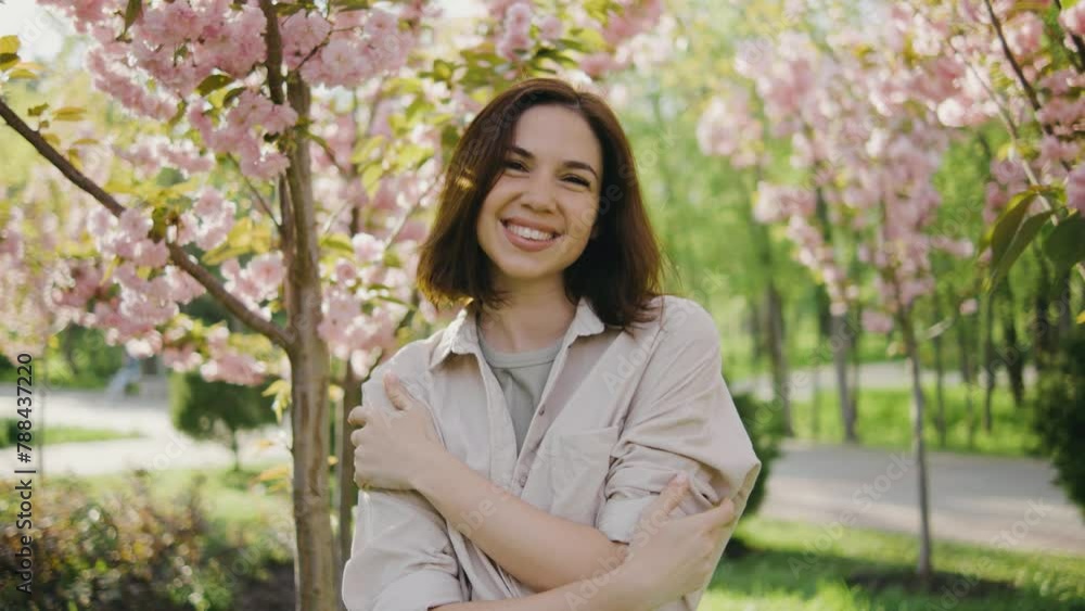 Portrait of a beautiful young woman. A girl walks in the park among pink blossoming sakura or cherry trees. Female smiles and looks at the camera against the background of pink spring trees on sunset Portrait of a beautiful young woman. A girl walks in the park among pink blossoming sakura or cherry trees. Female smiles and looks at the camera against the background of pink spring trees on sunset