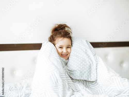 Happy little girl kid covered with blanket sitting on bed, looking at camera and laughs.