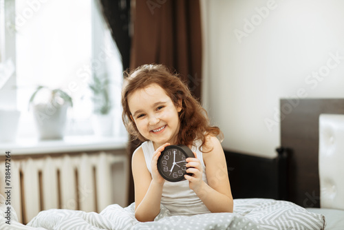 Cute little kid girl sitting on bed waking up in morning and holding alarm clock.