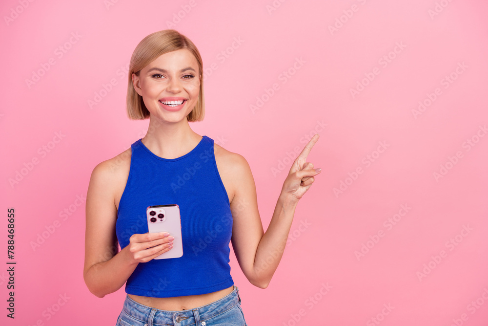 Photo of pretty young woman smart phone point finger empty space wear blue top isolated on pink color background Photo of pretty young woman smart phone point finger empty space wear blue top isolated on pink color background