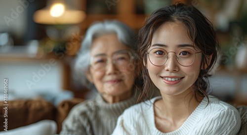 A young Asian woman in glasses smiling at the camera with an elderly Asian woman blurred in the background, indoors.
