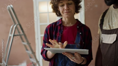 Cropped footage of Caucasian landlady with digital tablet presenting interior design ideas to faceless Black contractor while standing in renovating room and discussing future work photo