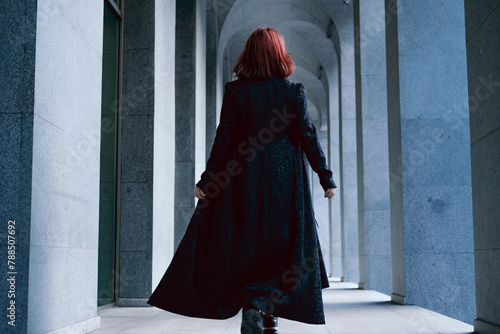 Young woman wearing long black cloak with red hair walking with her back turned in corridor between the columns of architectural building