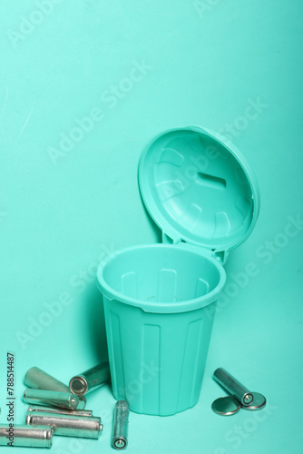 Used finger batteries near a miniature trash can. Light green background.