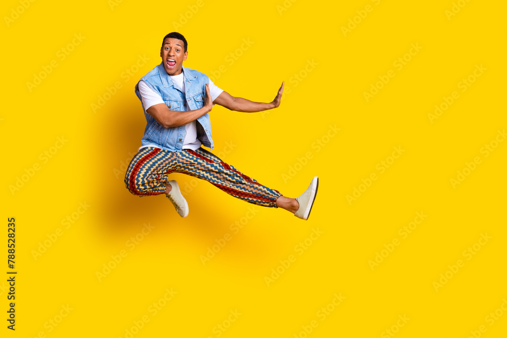 Full length photo of crazy eccentric guy dressed jeans waistcoat jumping near offer empty space isolated on yellow color background