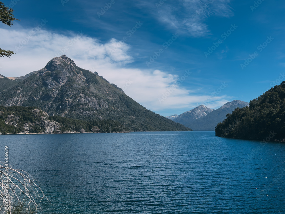 A beautiful lake with mountains in the background. The water is calm and clear. The sky is blue and there are some clouds. The mountains are covered in trees