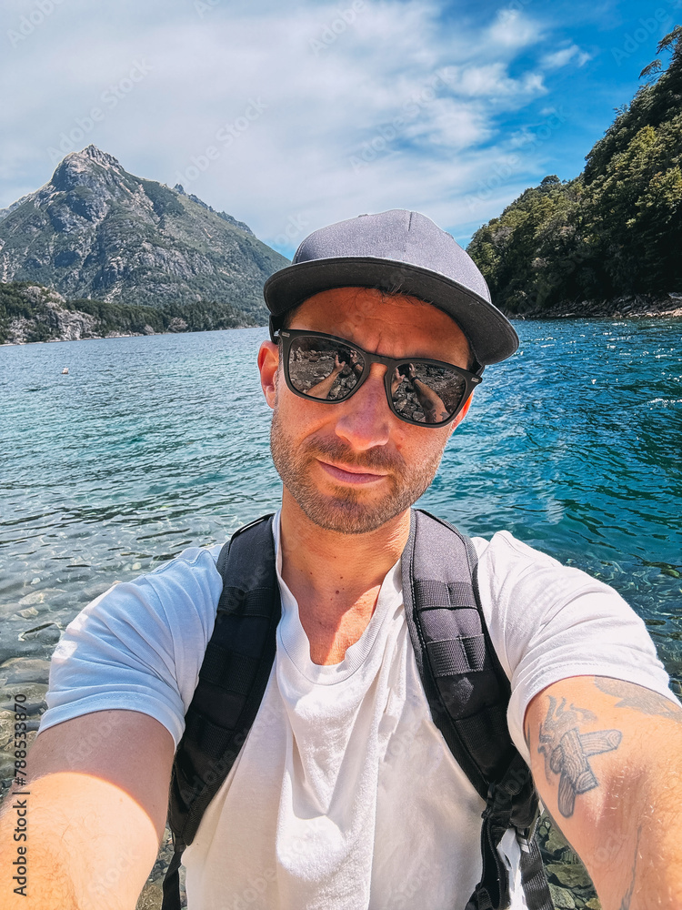 A man wearing sunglasses and a hat is taking a selfie in front of a body of water. Concept of relaxation and leisure, as the man is enjoying a day by the water