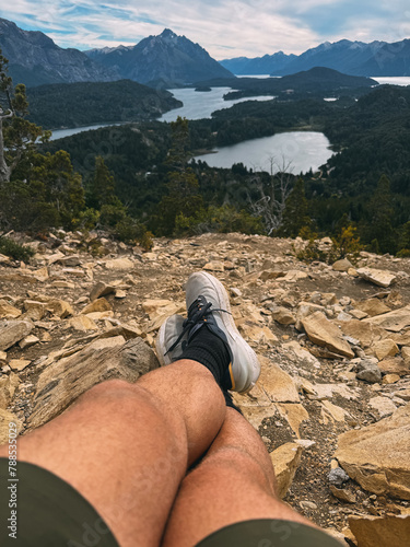 A man is laying on his back on a rocky hillside, with his legs crossed and his feet resting on his knees. The scene is peaceful and serene, with a beautiful view of the mountains