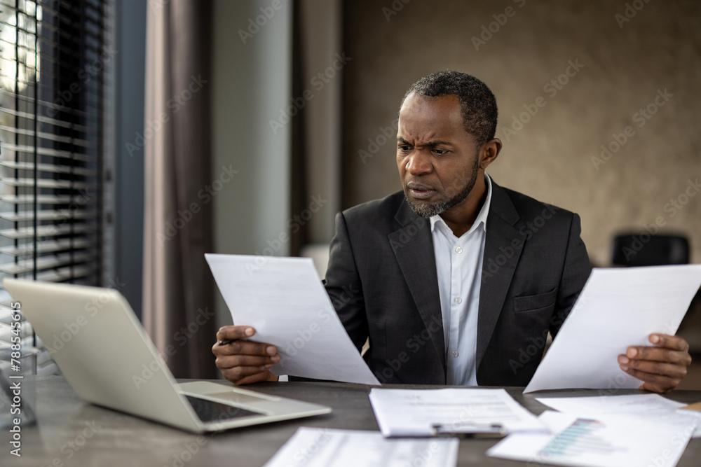 Sceptic formal male in business suit looking attentively on sheets of paper in both hands while sitting by desktop. Focused accountant comparing data on two documents from different departments.