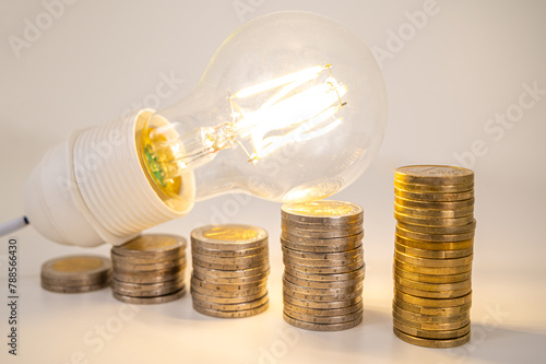 Light bulb lit, above stacks of coins. Increase in electricity tariffs, energy dependence, energy supplies.
