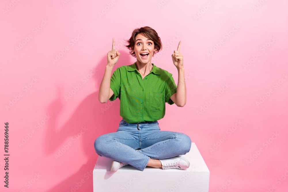 Fototapeta premium Full body photo of astonished pretty lady sit podium indicate fingers up empty space offer isolated on pink color background