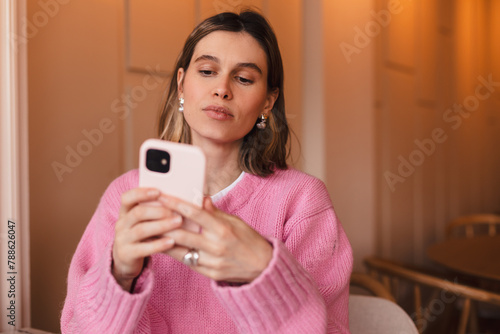 Young blonde woman using phone sits in cafe at table hold smartphone, answering texts, phone call, letters, posting photos, chatting or reading sms. Girl messaging, paying using phone.