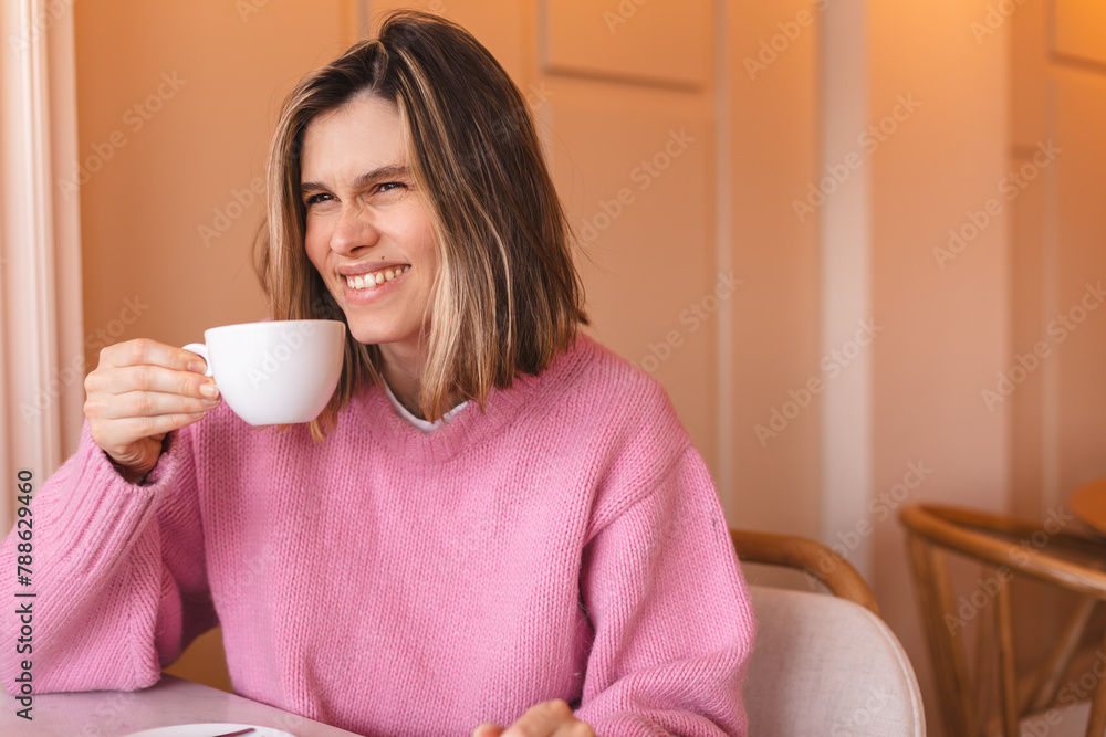 Portrait of joyful young woman enjoying a cup of tea and aroma at restaurant. Smiling pretty girl drinking hot coffee in cafe. Excited blonde woman drink cappuccino wearing pink sweater and laughing. Portrait of joyful young woman enjoying a cup of tea and aroma at restaurant. Smiling pretty girl drinking hot coffee in cafe. Excited blonde woman drink cappuccino wearing pink sweater and laughing.