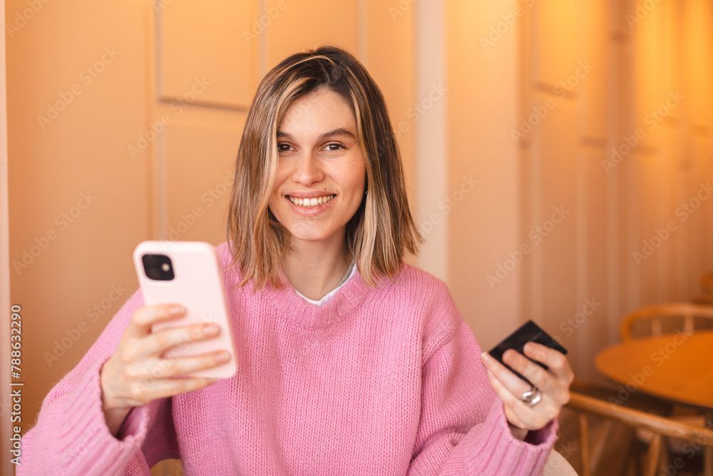 Happy young blonde woman customer shopper holding credit card using cell phone mobile app buying fashion clothes paying online make purchase in ecommerce digital store on smartphone in cafe. Happy young blonde woman customer shopper holding credit card using cell phone mobile app buying fashion clothes paying online make purchase in ecommerce digital store on smartphone in cafe.