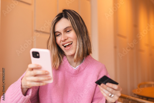 Happy young blonde woman customer shopper holding credit card using cell phone mobile app buying fashion clothes paying online make purchase in ecommerce digital store on smartphone in cafe.