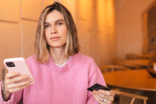 Calm young woman wear pink knit sweater clothes with credit card in hand using mobile phone paying for purchase online relaxing sitting at table in cafe. Serious blonde girl.