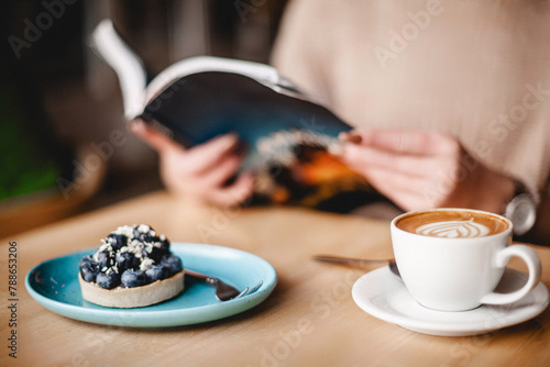 A woman relishes a tranquil moment, engrossed in a book while savoring a blueberry tart and coffee. The scene exudes relaxation and leisure, a delightful blend of literature and culinary indulgence.