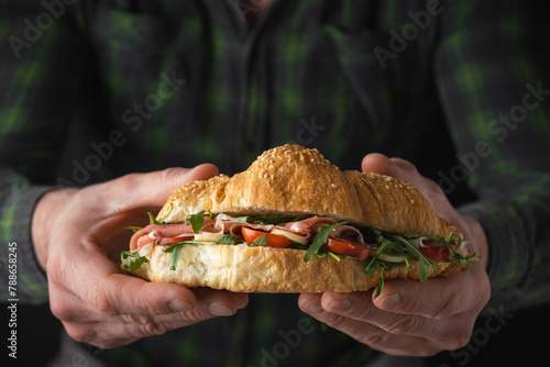 Man wearing checkered shirt and holding freshly baked croissant with iberian ham, tomatoes and salad filling. Close up