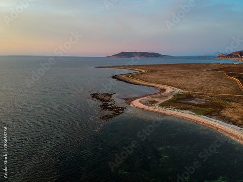 recorte da costa de cor castanha deserta a entrar no mar com uma ilha ao fundo, céu azul sem nuvens