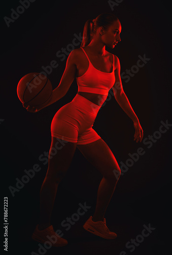 A beautiful athletic blonde girl in white shorts and a top plays basketball on a black background in red light.