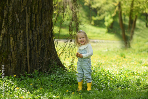 A beautiful little girl walks in a field of dandelions. Child's spring walks. A two-year-old girl with blond hair wearing yellow rubber boots