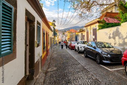 Tourists walk down the colorful Rua de Santa Maria street towards the Art of Open Doors district of shops and cafes in the Zona Velha old town of Funchal, Portugal on the Canary island of Madeira.