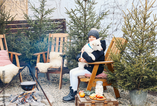 Smiling teenage girl in warm clothes holding cute white puppy in arms while sitting in backyard surraunded with coniferous trees, hugging dog and enjoying winter weather in company of best pet friend photo