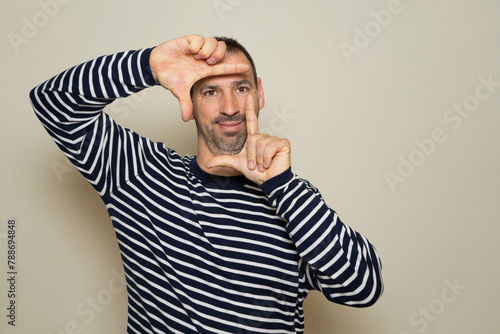A happy bearded man blinks his eyes and looks through a frame formed by his hands. He makes a frame with his fingers, isolated on beige background with copy space for text. People concept.