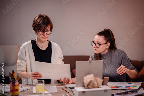 Two focused women engage in collaborative work, one sketching ideas with the other providing input, in a warmly lit office environment