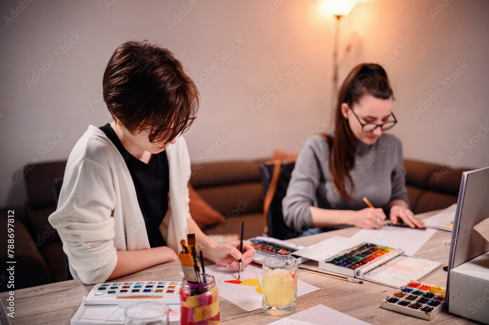 Two concentrated women involved in an artistic project, with one painting and the other measuring, surrounded by creative tools