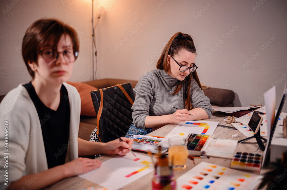 Fototapeta premium Two artists intently craft a color narrative, one with brush in hand, the other with pen poised, against the backdrop of a warm studio light.
