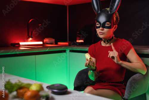 beautiful girl in a bdsm rabbit mask and a bright red dress eats an apple in the kitchen in neon light promoting a healthy lifestyle vegetarianism