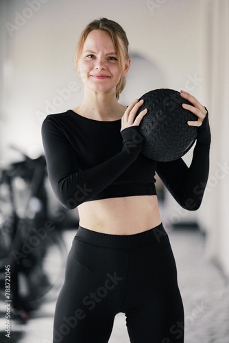 Fotografie With a confident smile, a woman holds a medicine ball at chest height, exuding fitness and poise in her gym attire