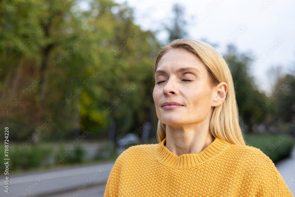A middle-aged woman with blonde hair enjoys the warm sunlight in a serene park setting, wearing a cozy yellow sweater, eyes closed, embracing tranquility.