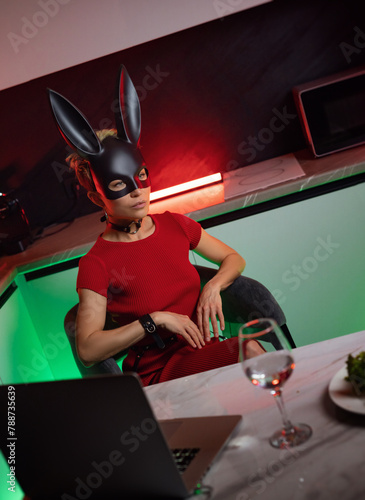 A beautiful girl in a bdsm-style rabbit mask and a bright red dress with leather straps is posing sweetly in the kitchen in neon light
