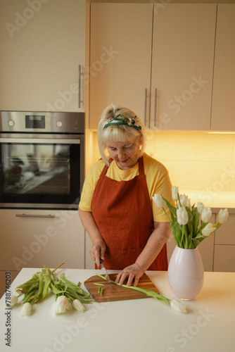 Happy Caucasian 30s woman taking care of cut fresh bouquet of tulips in kitchen. Housewife hands of woman cutting flower on wooden board and putting fresh tulips into the vase.