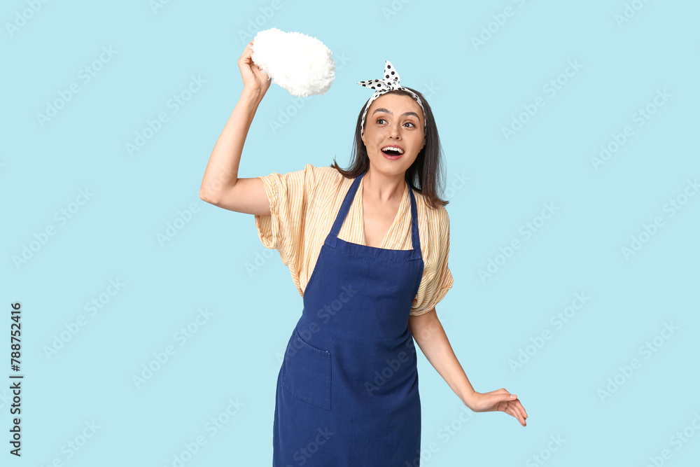 Portrait of happy young housewife in apron with pp duster on blue background Portrait of happy young housewife in apron with pp duster on blue background