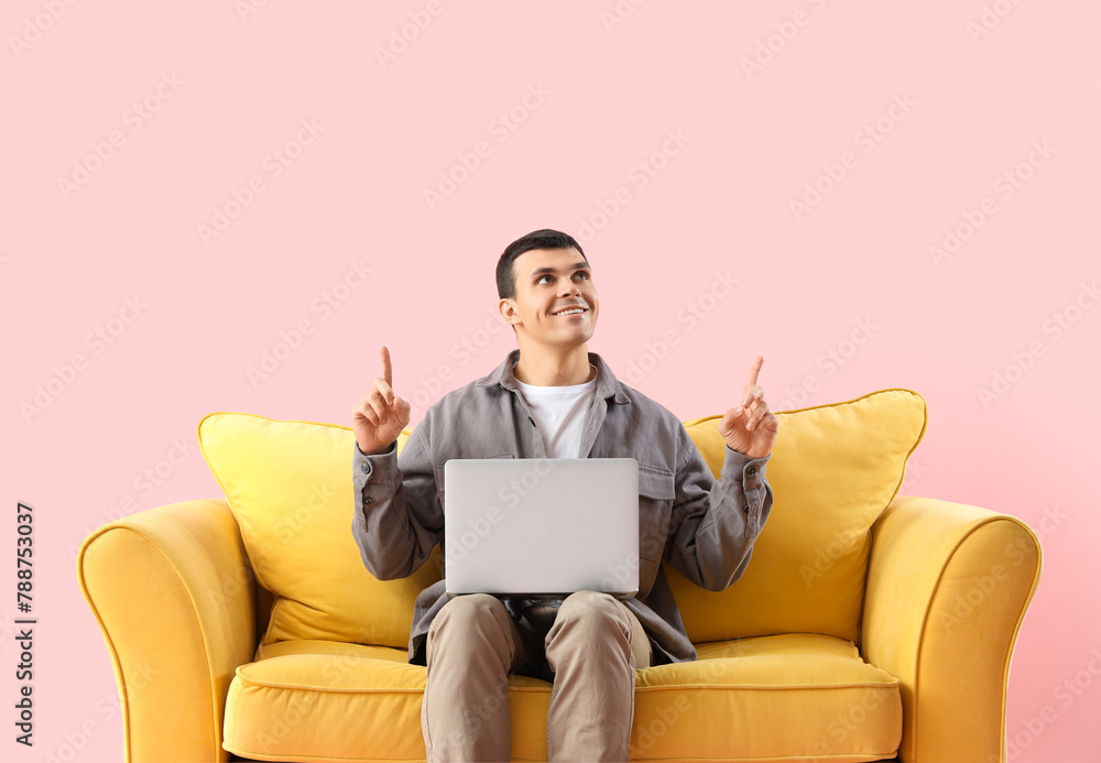 Young man with laptop on sofa pointing at something against pink background Young man with laptop on sofa pointing at something against pink background