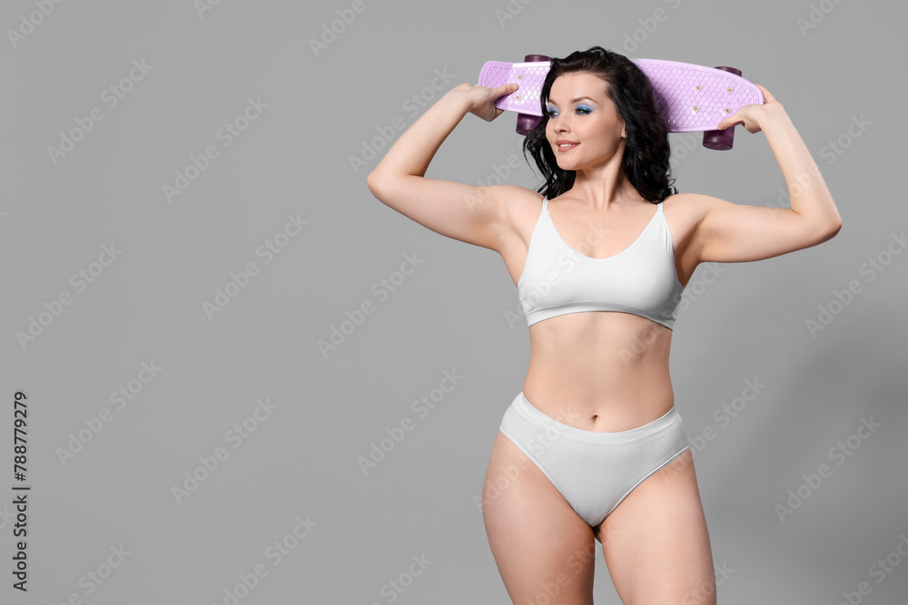 Fototapeta premium Beautiful young body positive woman with skateboard in stylish underwear on grey background